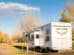 A large white RV with "X-Aire" branding is parked on a paved area surrounded by autumn trees with yellow leaves. The sky is clear with a few scattered clouds.