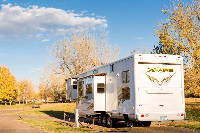 A large white RV with "X-Aire" branding is parked on a paved area surrounded by autumn trees with yellow leaves. The sky is clear with a few scattered clouds.
