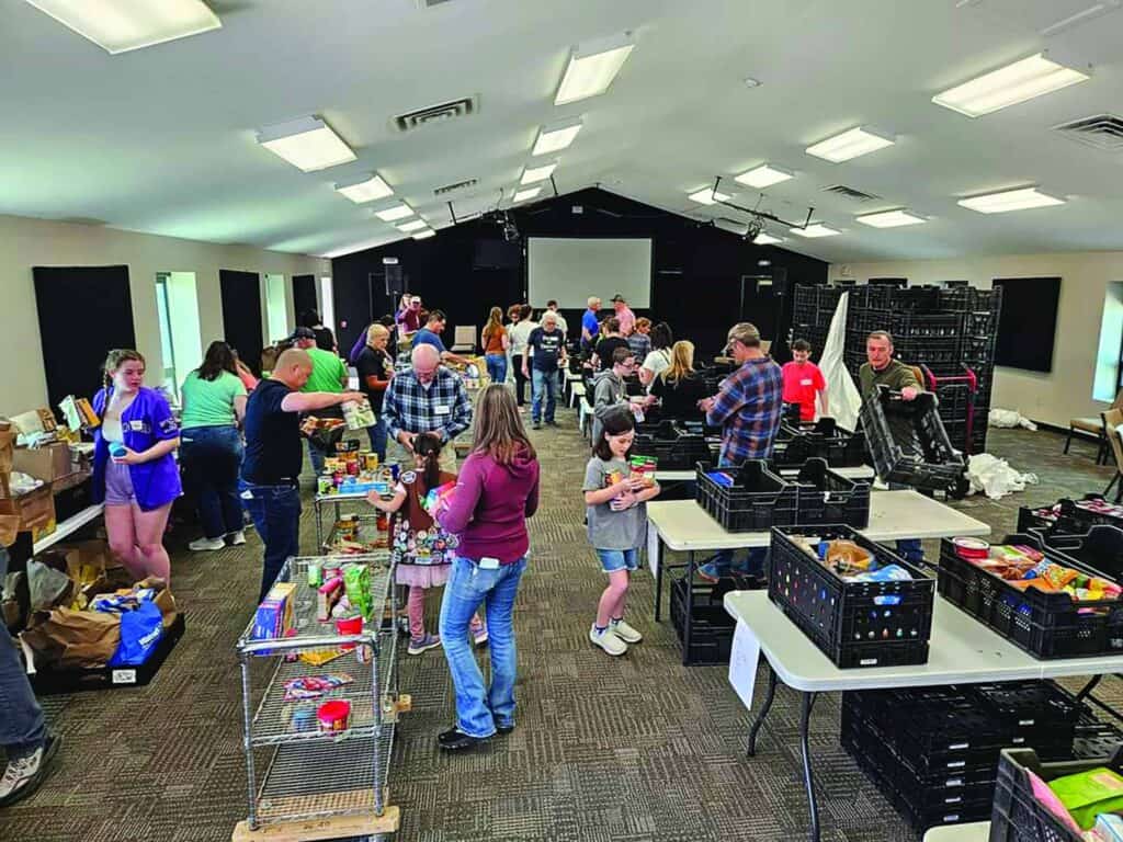A group of people of various ages packing food donations in a large, well-lit room with tables filled with groceries and black crates.