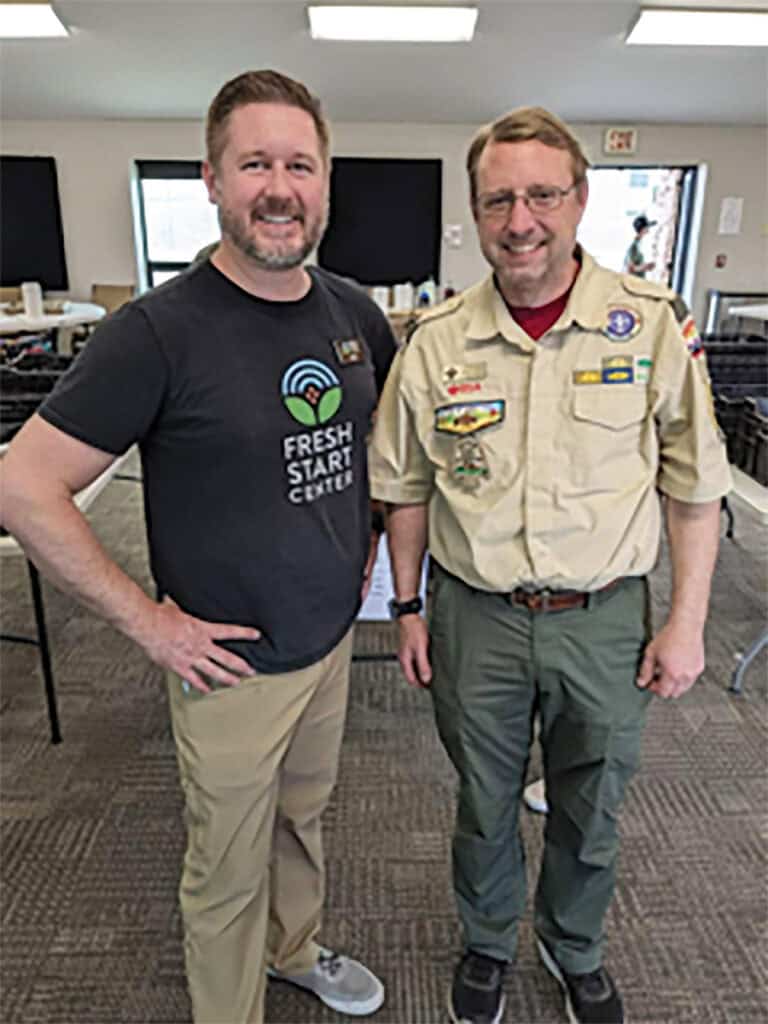 Two men smiling indoors, one in a dark t-shirt and beige pants, the other in a boy scout uniform with badges.