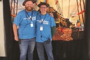 Two individuals in matching blue shirts and hats smiling for a photo in front of a nautical-themed backdrop.