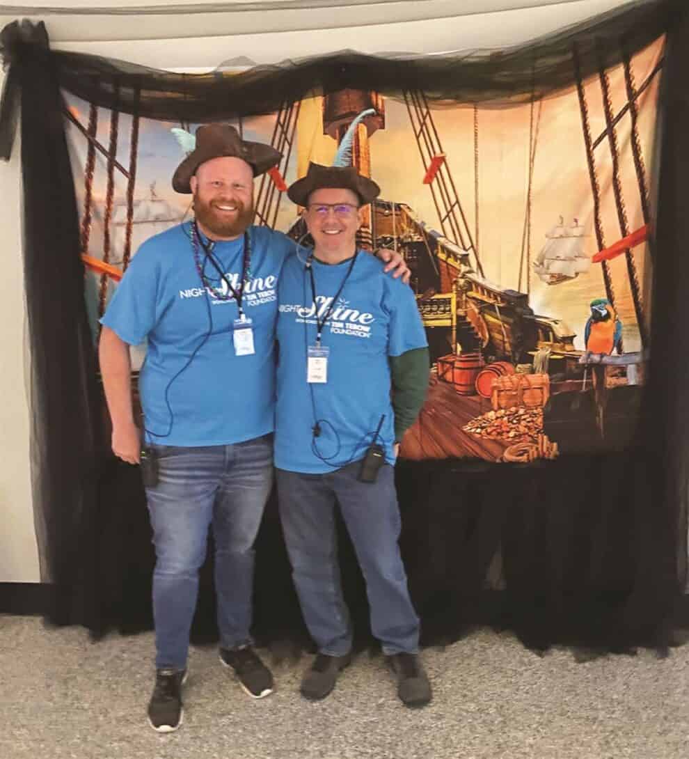 Two individuals in matching blue shirts and hats smiling for a photo in front of a nautical-themed backdrop.