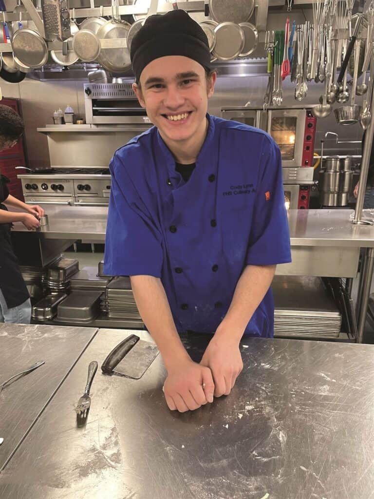 A smiling young chef in a blue uniform stands at a stainless steel countertop in a kitchen.