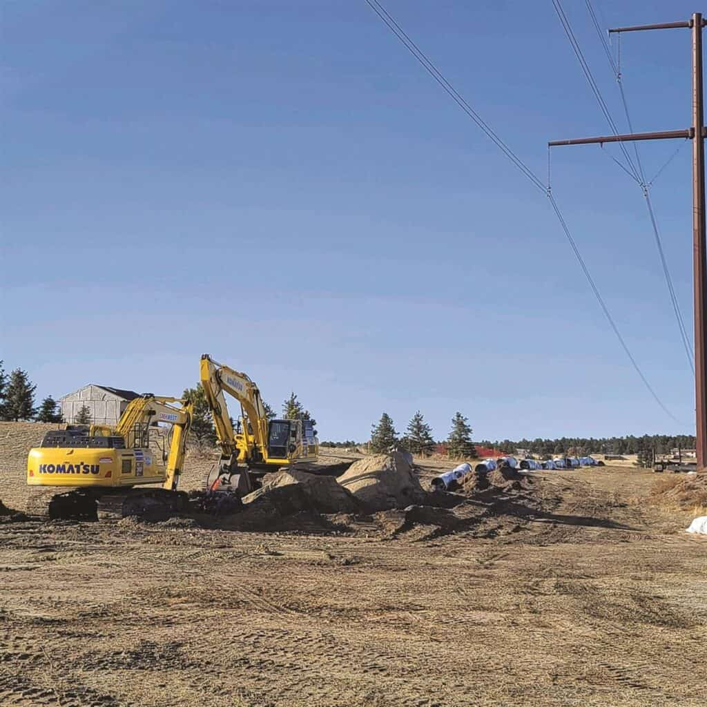 Construction equipment and workers on a dirt site with utility poles in the background.