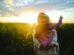 A child on an adult's shoulders points at a sunset over a lush field.