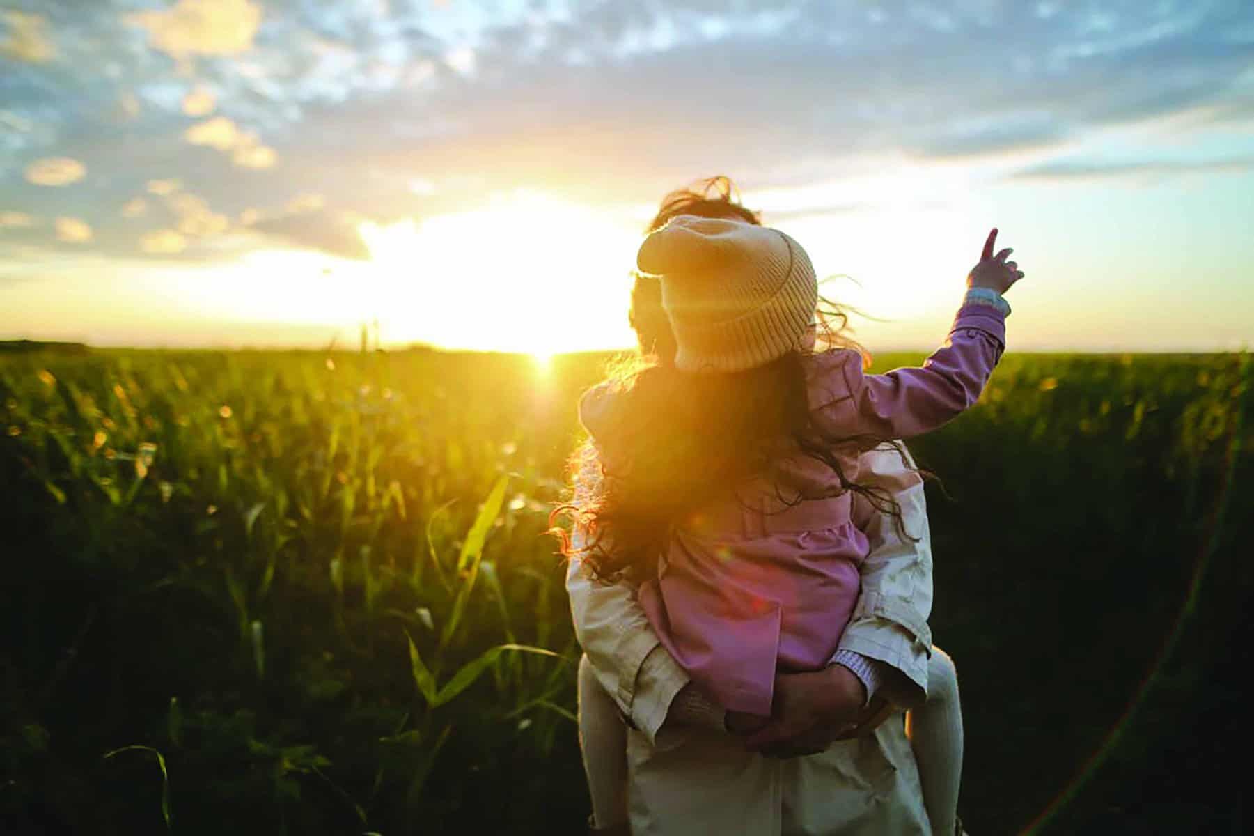 A child on an adult's shoulders points at a sunset over a lush field.