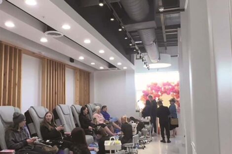A row of people seated in massage chairs receiving pedicures in a modern, brightly lit nail salon with high ceilings and a white interior.