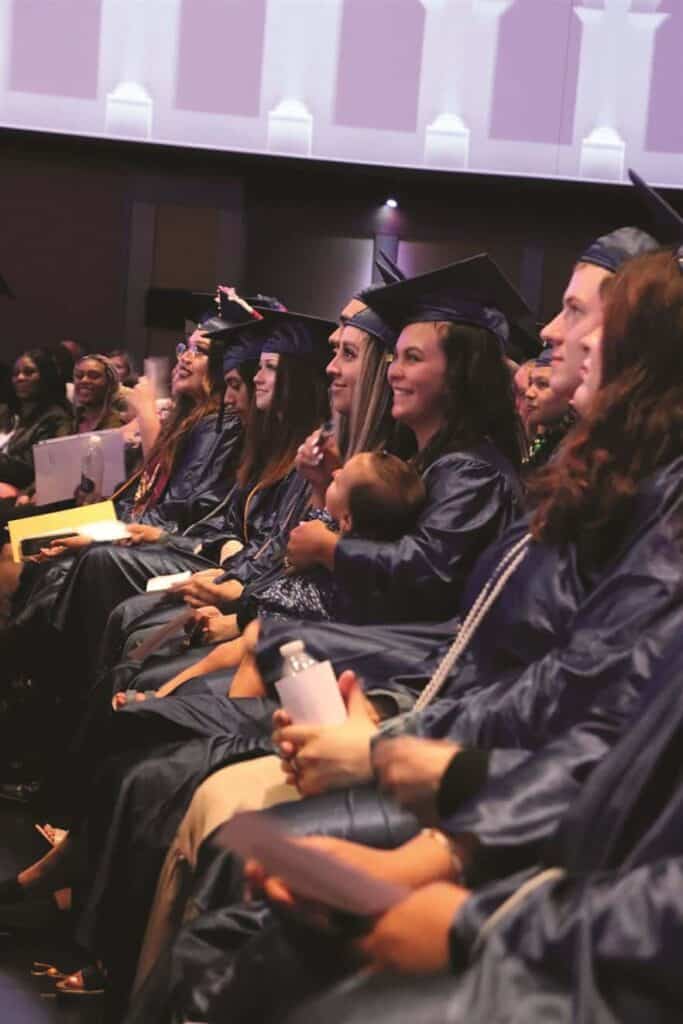 A group of graduates in caps and gowns sit in rows, smiling and holding diplomas, during a ceremony in an indoor venue.