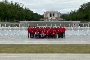 A group of people wearing red jackets poses in front of the world war ii memorial fountains with the lincoln memorial in the background.