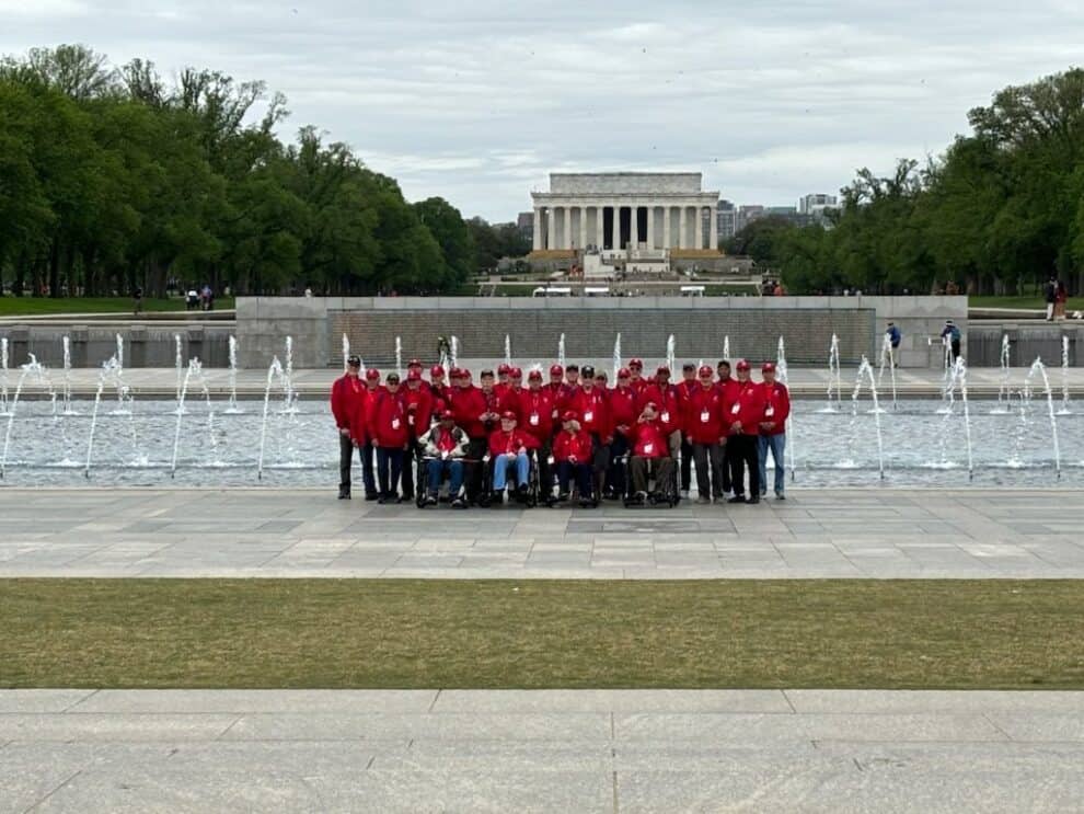 A group of people wearing red jackets poses in front of the world war ii memorial fountains with the lincoln memorial in the background.