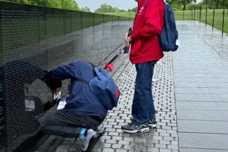 Two men at the vietnam veterans memorial, one crouching to touch the wall, with the washington monument in the background.