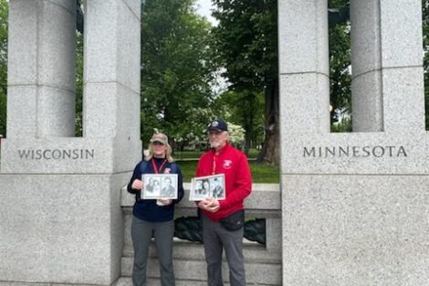 Two people stand between columns marked "wisconsin" and "minnesota" at a memorial, each holding a picture frame.