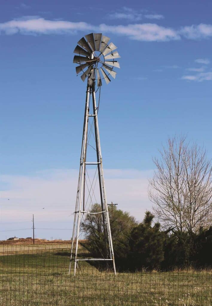 A tall metal windmill stands in a grassy field with a clear blue sky and sparse trees in the background.