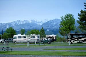Several motorhomes parked in a campsite with picnic tables in the foreground and snow-capped mountains in the background.