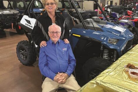 Vernon & Barb Clark pose for a photo in a showroom filled with ATVs and motorcycles. The man is seated, wearing a blue shirt, while the woman stands behind him with her hands on his shoulders.