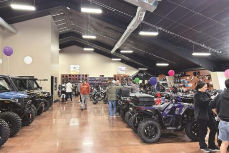 A showroom with motorcycles, ATVs, and people browsing. Balloons are attached to various vehicles. The ceiling is high with exposed ducts, and the floor has a polished finish.