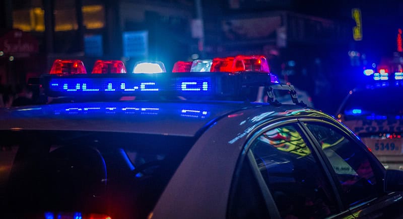Police car with flashing blue and red lights at night. Other vehicles with flashing lights are visible in the background.