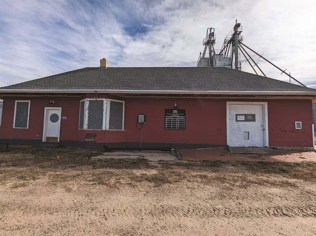 A red, single-story building with a gray roof and several windows, located in a rural area. Industrial structures are visible in the background. The ground in front is barren and covered in dirt.