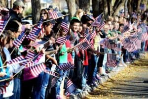 A large group of people, including children, standing in a line along a sidewalk, holding small American flags. Trees line the street in the background.