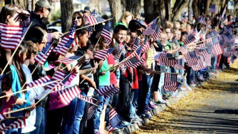 A large group of people, including children, standing in a line along a sidewalk, holding small American flags. Trees line the street in the background.