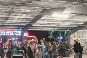 People dancing under a metal-roofed pavilion, with a country band performing onstage in the background. Strings of lights and colorful stage lighting add to the lively atmosphere.