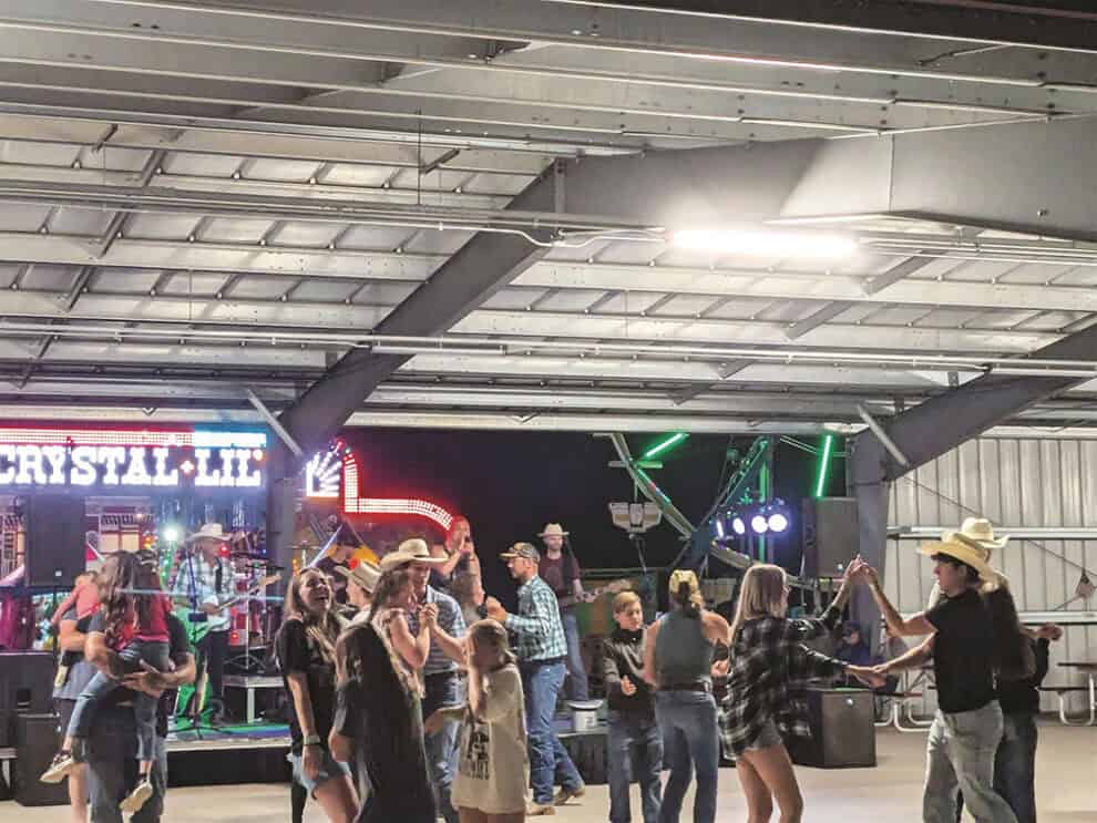 People dancing under a metal-roofed pavilion, with a country band performing onstage in the background. Strings of lights and colorful stage lighting add to the lively atmosphere.