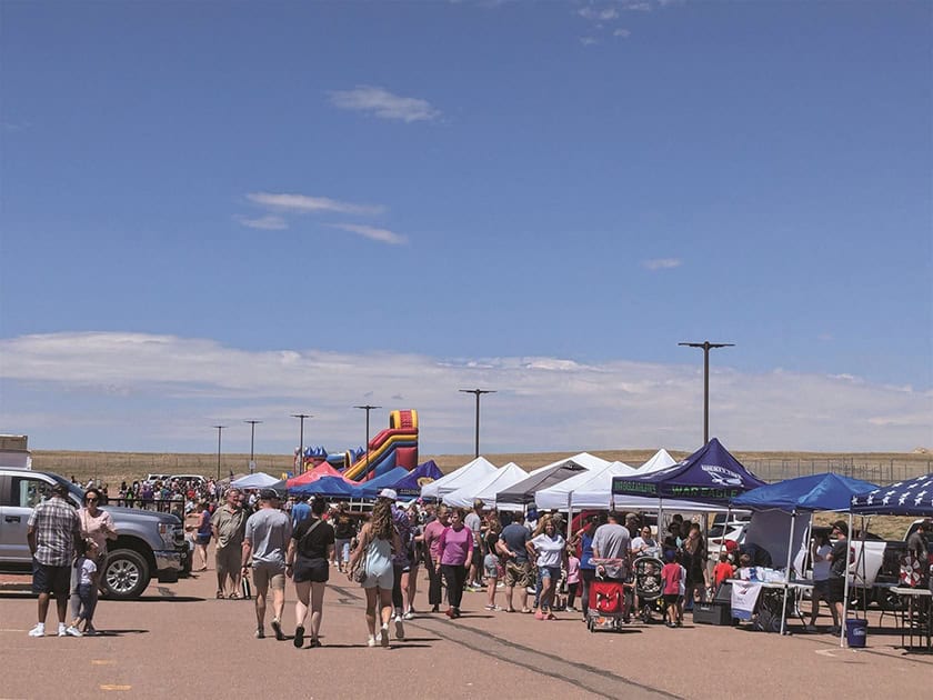 Outdoor fair with people walking between vendor tents and a colorful inflatable slide, under a clear blue sky.