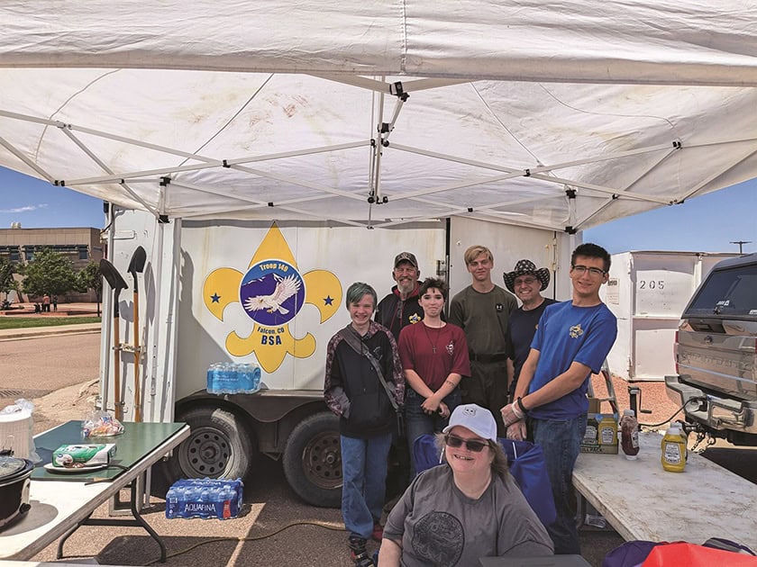 A group of people stands under a white canopy with a Boy Scouts of America emblem on a backdrop. Camping gear, tables, and a barbecue setup are visible. They appear to be in an outdoor setting.