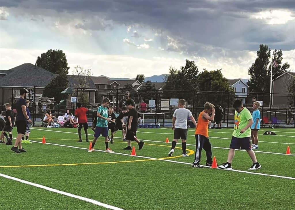 Children participating in outdoor sports activities on a grassy field with traffic cones, under a cloudy sky, with houses and trees in the background.