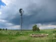 A tall windmill stands in a lush green field, with a cloudy sky and rain in the distance. An empty stone circular foundation is visible in the foreground.