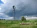 A tall windmill stands in a lush green field, with a cloudy sky and rain in the distance. An empty stone circular foundation is visible in the foreground.