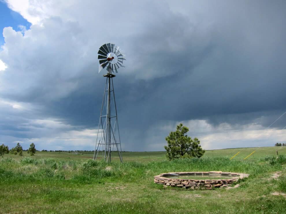 A tall windmill stands in a lush green field, with a cloudy sky and rain in the distance. An empty stone circular foundation is visible in the foreground.