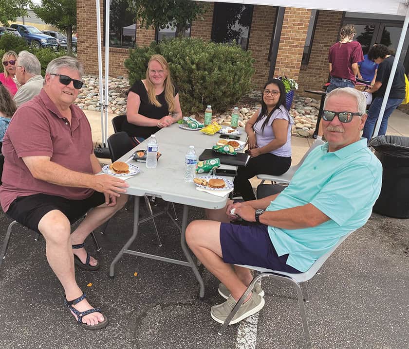 Four people sit around a table outdoors, eating burgers and chips. Other people and a brick building are in the background.