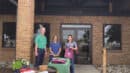 Three individuals standing in front of the Farmers State Bank building. A table with promotional materials and a grill is set up on the sidewalk.