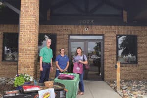 Three individuals standing in front of the Farmers State Bank building. A table with promotional materials and a grill is set up on the sidewalk.