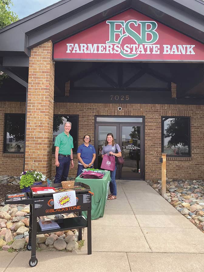 Three individuals standing in front of the Farmers State Bank building. A table with promotional materials and a grill is set up on the sidewalk.