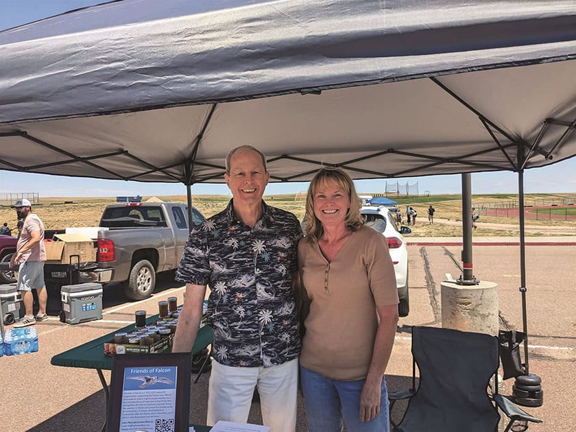 Two people standing under a tent at an outdoor event. The man is wearing a patterned shirt and the woman a brown top. They are in front of a table with informational materials.