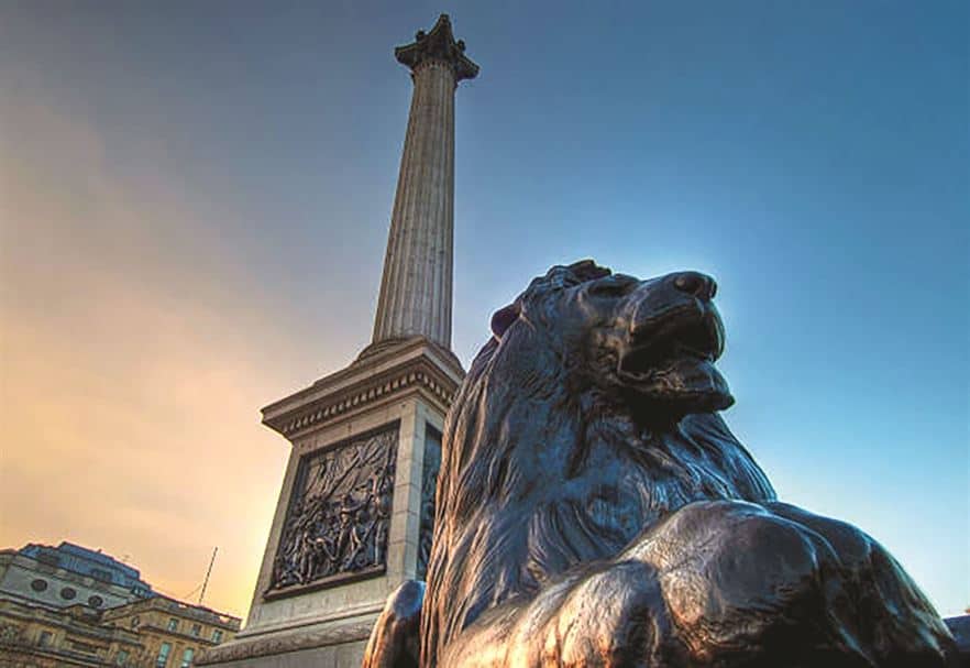 A view of Nelson's Column with a bronze lion sculpture in the foreground, located in Trafalgar Square, London, under a clear sky.