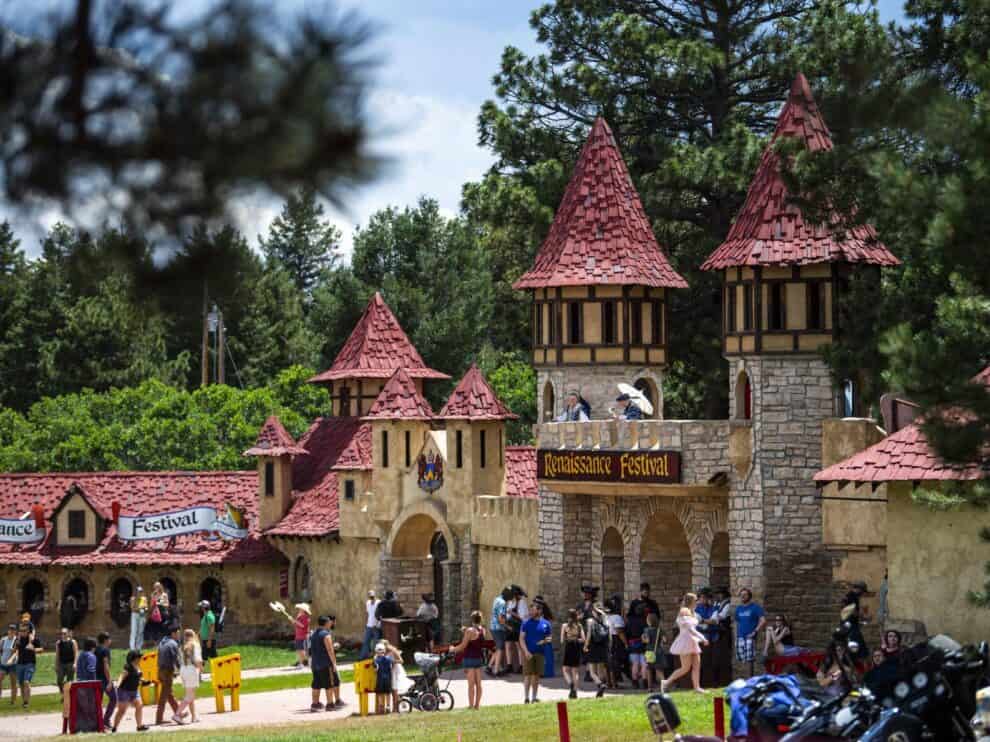 People gather outside a medieval-style castle with red roofs at a Renaissance Festival, surrounded by trees and various stalls.