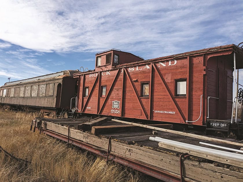 A rusty red train caboose labeled "Rock Island" and an old passenger car sit on abandoned tracks in a grassy field under a partly cloudy sky.