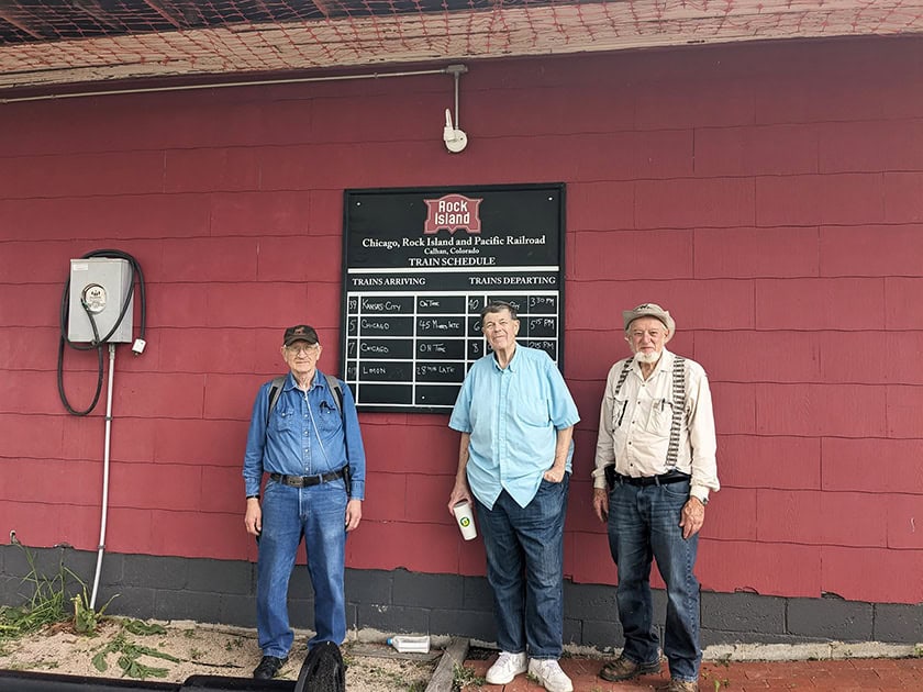 Three elderly men stand in front of a bulletin board labeled "Chicago, Rock Island and Pacific Railroad Train Schedules." The backdrop is a red wall and there is a railing in front of them.