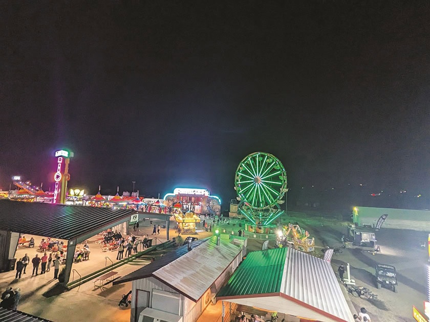 Nighttime carnival scene featuring a lit Ferris wheel, colorful tents, illuminated rides, and scattered groups of people. The area is surrounded by dark skies.