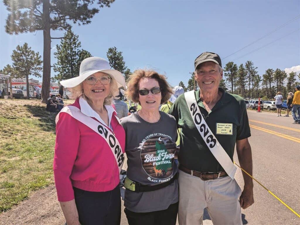 Three people stand outdoors at a festival, smiling at the camera. Two people wear "2023" sashes, and one wears a festival T-shirt. Trees and festival activities are in the background.