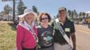 Three people stand outdoors at a festival, smiling at the camera. Two people wear "2023" sashes, and one wears a festival T-shirt. Trees and festival activities are in the background.