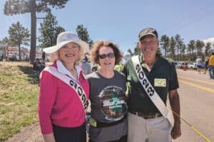 Three people stand outdoors at a festival, smiling at the camera. Two people wear "2023" sashes, and one wears a festival T-shirt. Trees and festival activities are in the background.