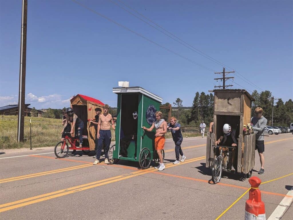 Participants push and ride mobile outhouse structures along a road in a race event under a clear sky.