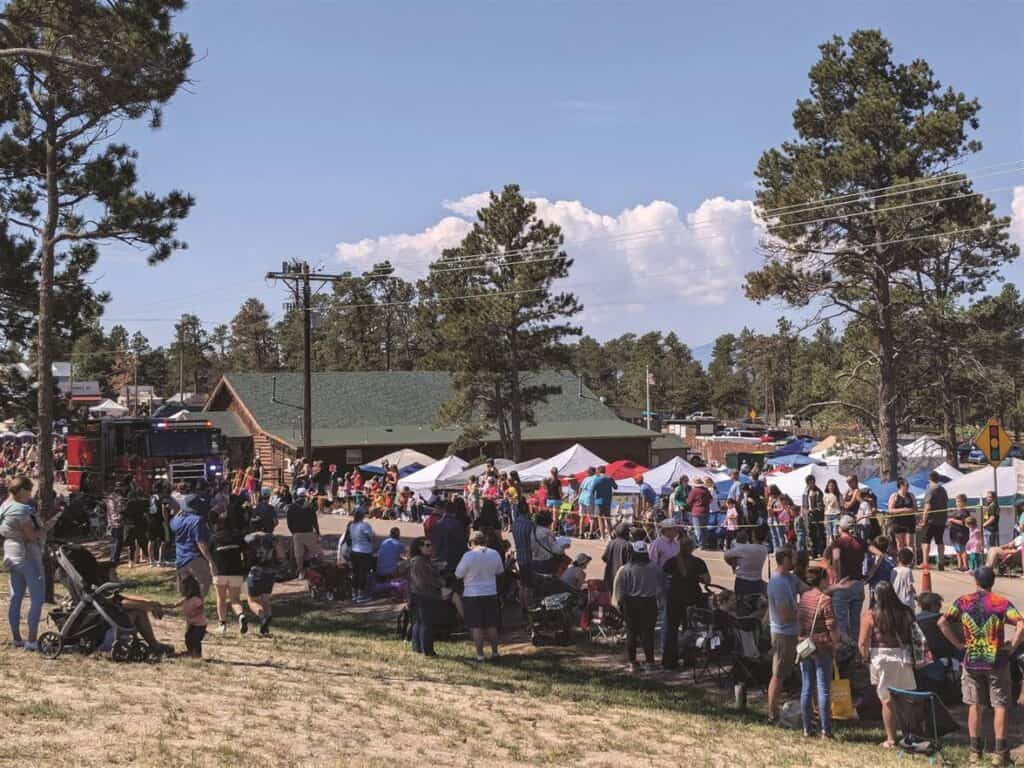 Outdoor festival with people gathered around tents and booths in a tree-filled area on a clear day.