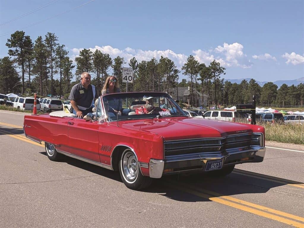 A red classic convertible car with three people driving on a road during daylight, surrounded by trees and a speed limit sign in the background.