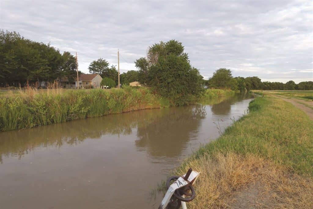 A narrow canal flows through a rural landscape with houses and trees on the left bank, and grassy fields on the right. A cloudy sky looms overhead.