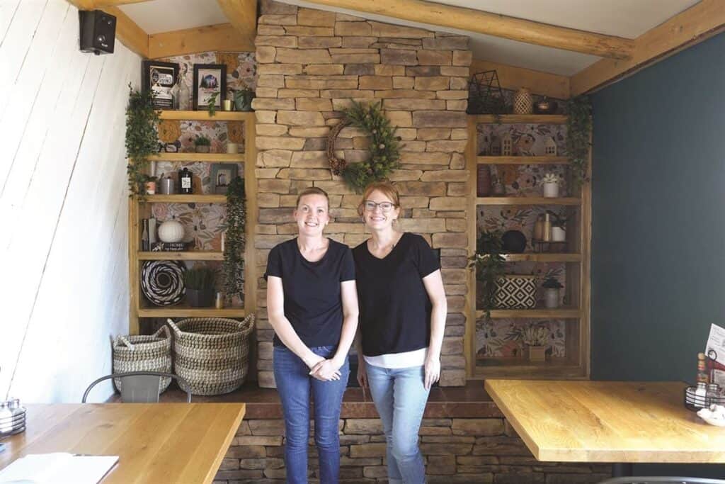 Two women stand smiling in front of a stone fireplace with wooden shelves on either side, decorated with various ornaments, in a cozy indoor setting.